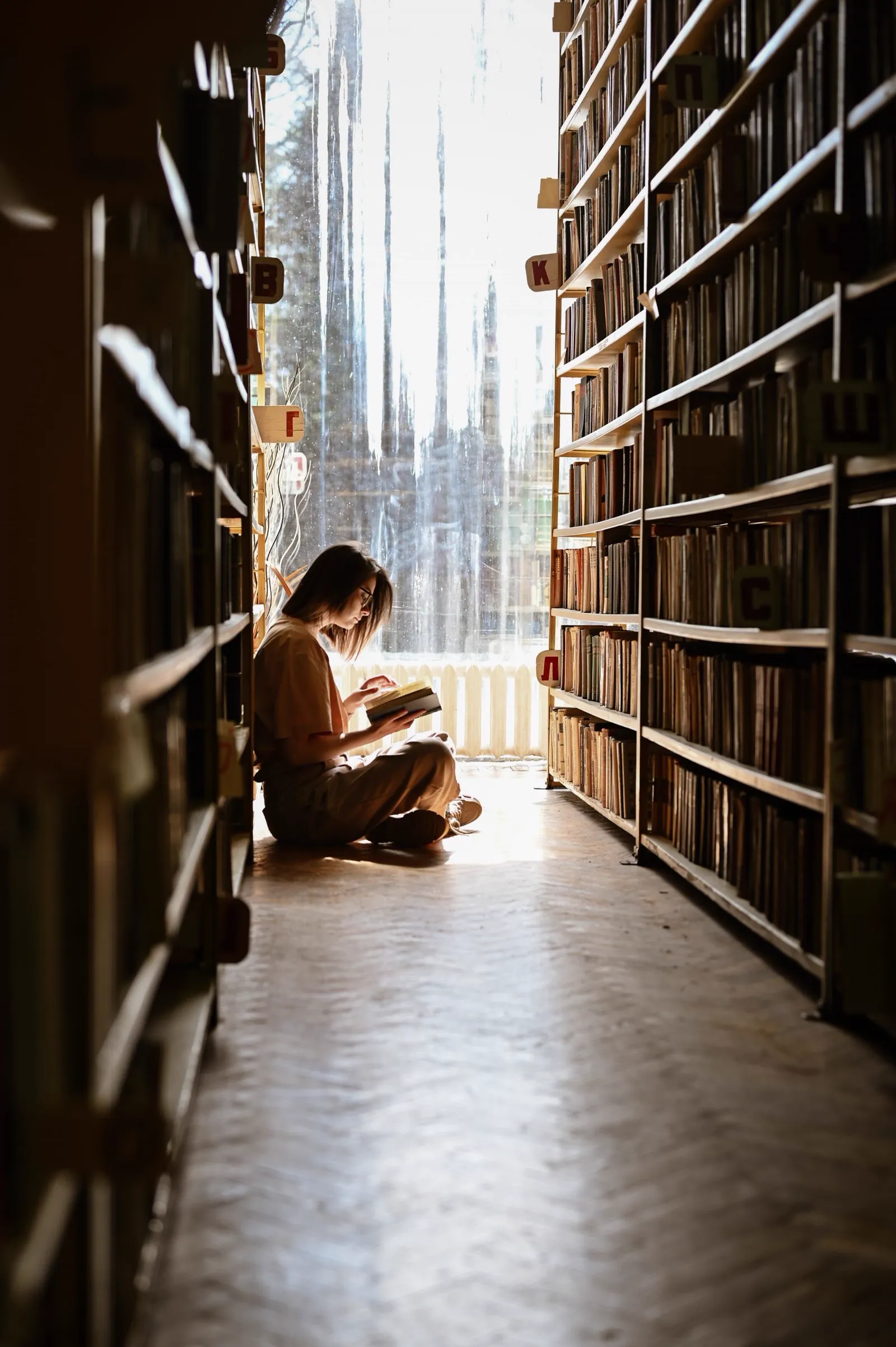 A girl reads on the floor of a sunlit library aisle